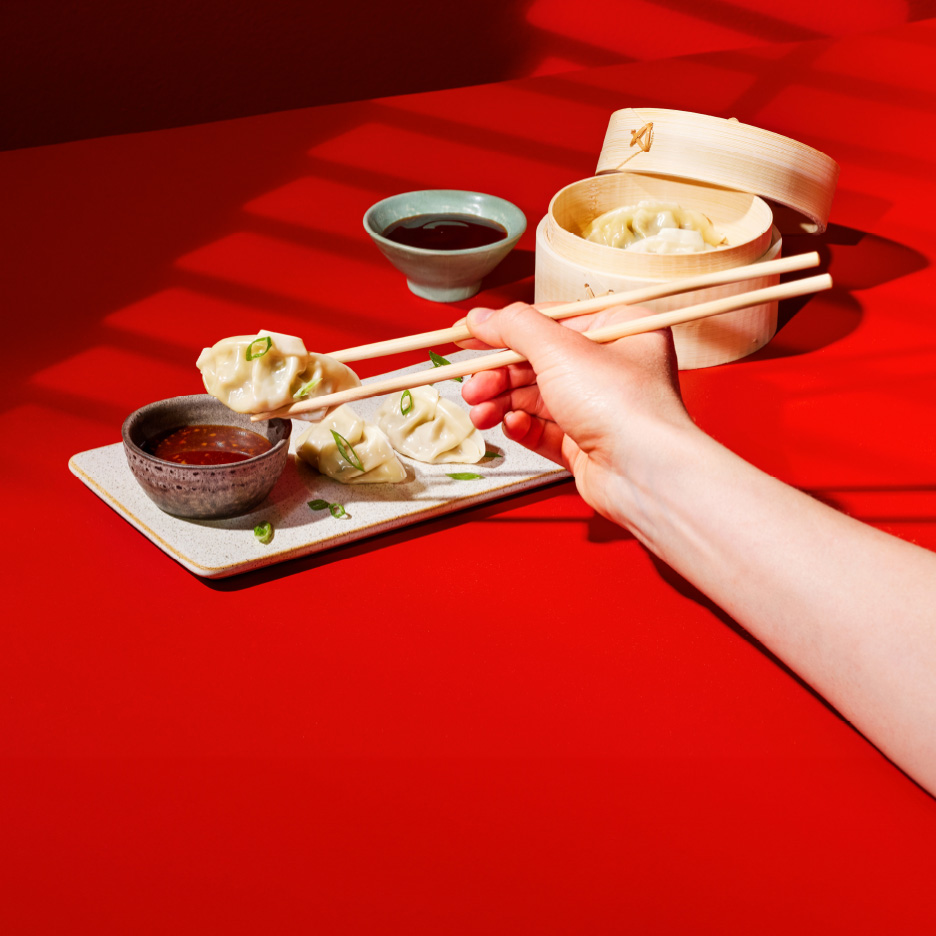 En hånd holder spisepinde med en dumpling over en skål sauce, med flere dumplings på en rød baggrund.