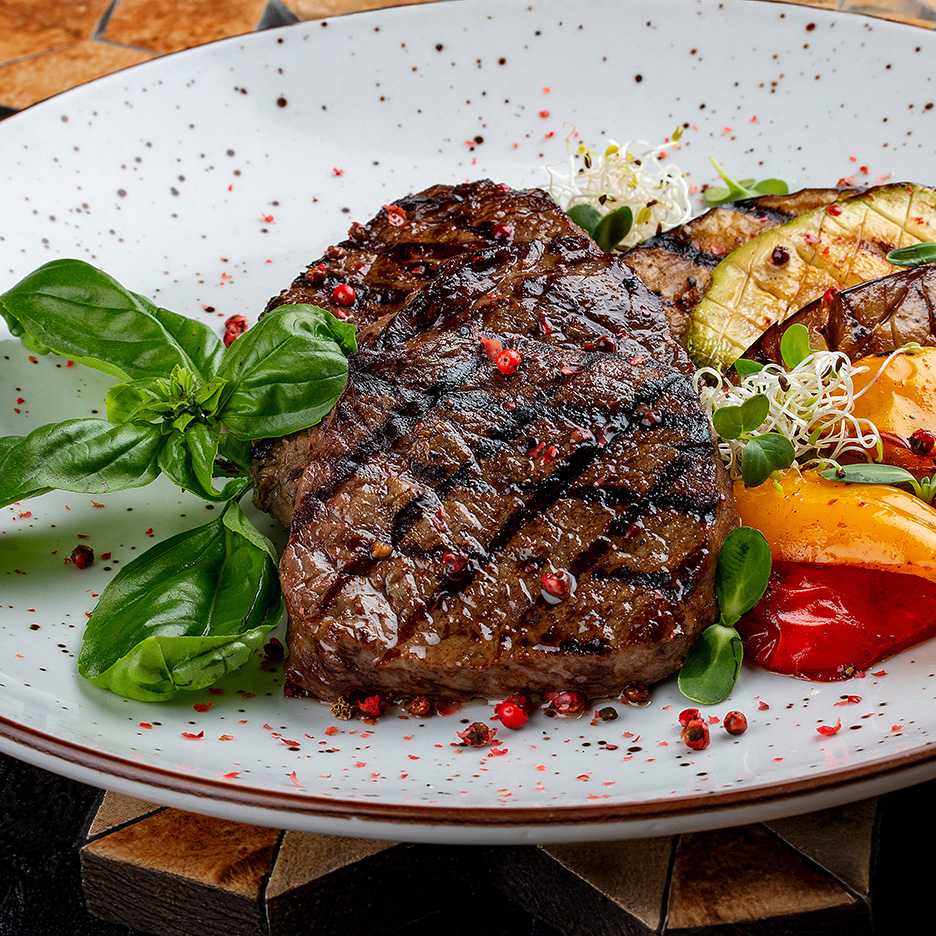 Grillet bøf med grøntsager, basilikum og rød peber på en hvid tallerken.