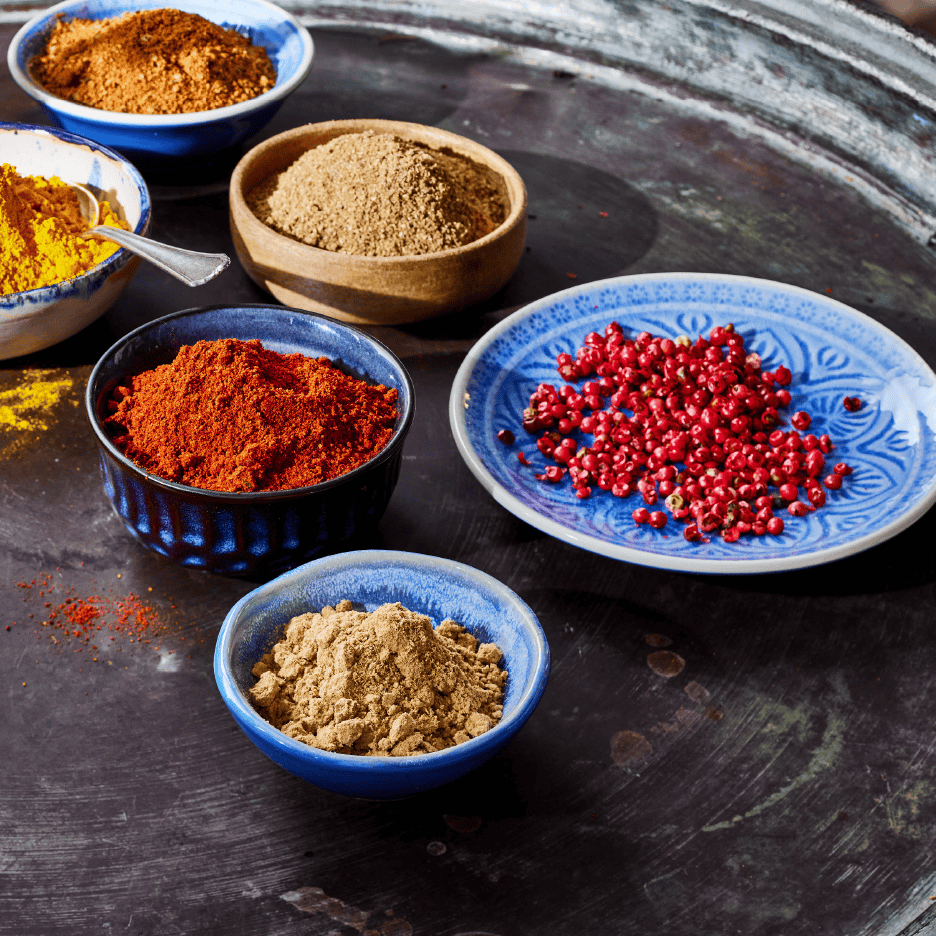 Forskellige krydderier i skåle, herunder paprika, gurkemeje og lyserøde peberkorn.