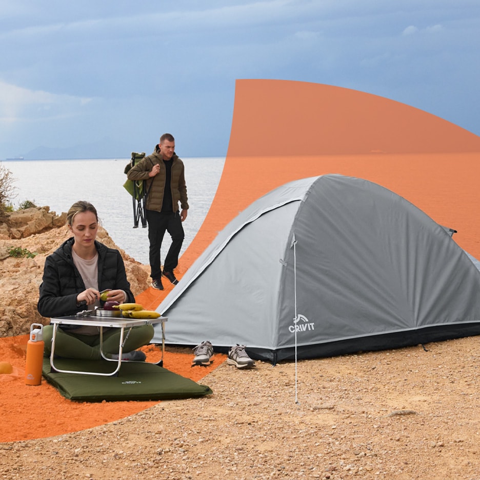 Campingtelt og campingudstyr ved havet med en kvinde, der tilbereder mad, og en mand, der går.