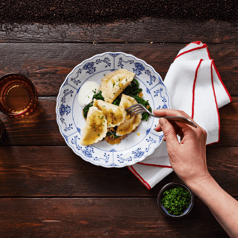 En person spiser pierogi med spinat og creme fraiche fra en tallerken på et træbord.