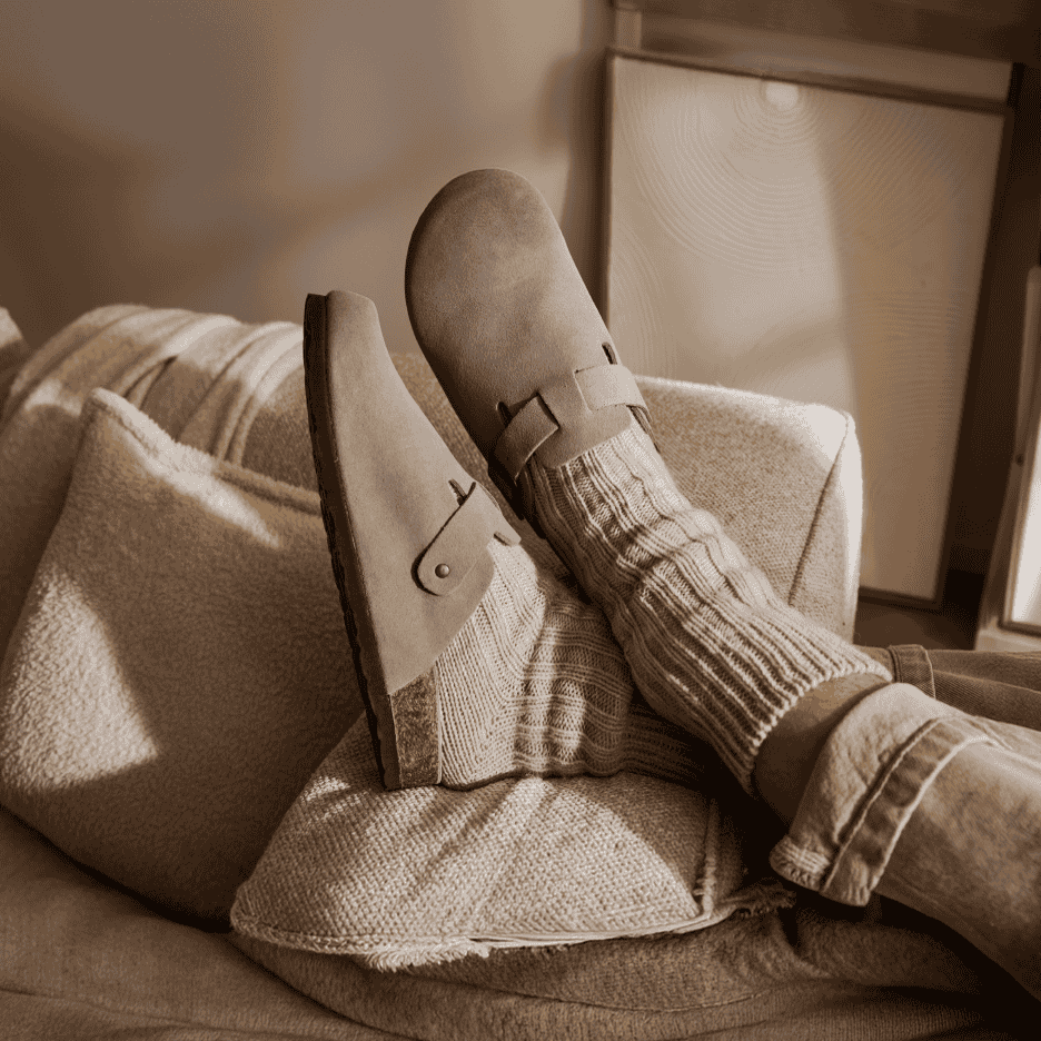 Fødder i hjemmesko og strikkede sokker hviler på en sofa med puder.