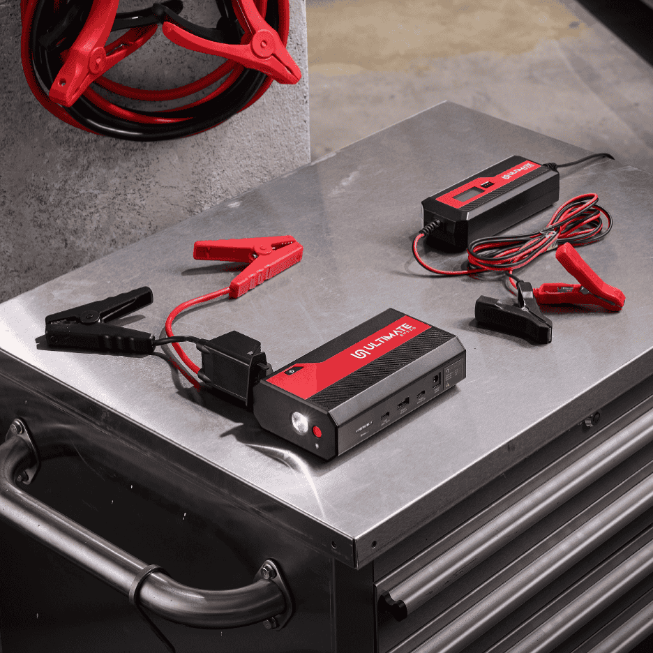 Jumpstarter og batterioplader på et metalbord med startkabler i baggrunden.
