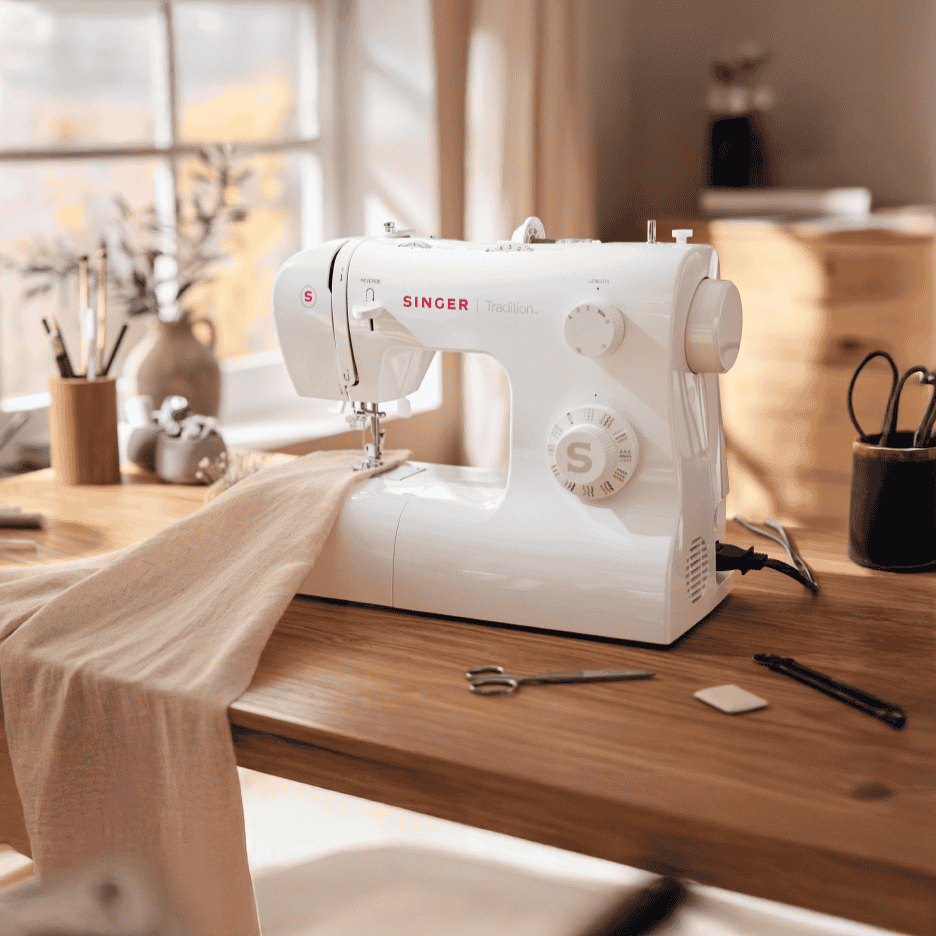 Hvid symaskine på et træbord med stof og sytilbehør i et lyst rum.