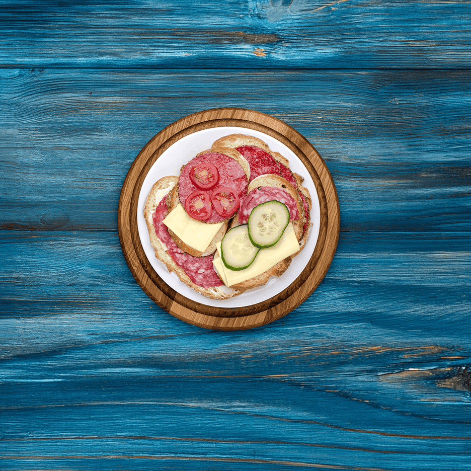 En tallerken med rugbrødsmadder med salami, ost, agurk og tomat på et blåt træbord.