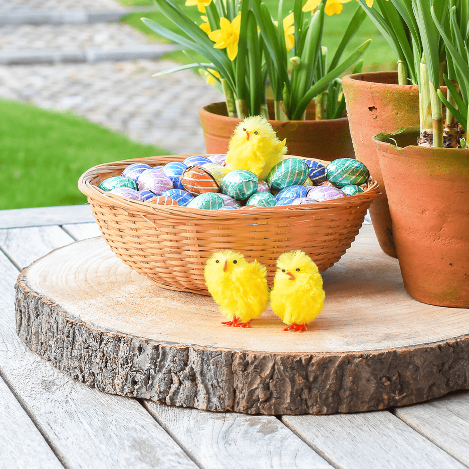 Påskepynt med en kurv fyldt med chokoladeæg, gule kyllinger og påskeliljer i potter.