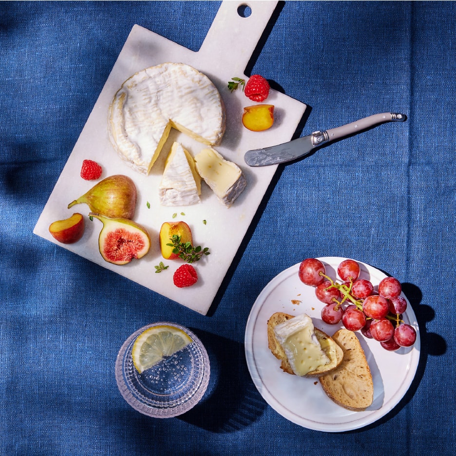 Ostebord med brie, figner, hindbær, nektariner og druer på et blåt klæde.