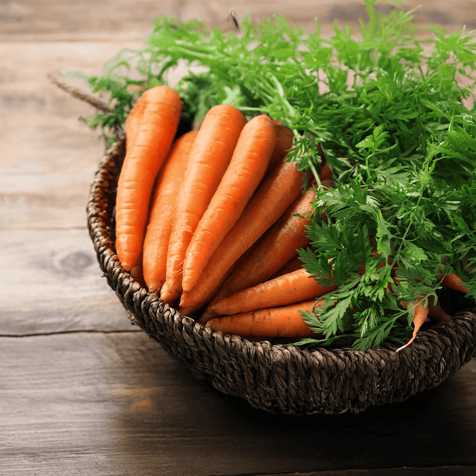 Friske gulerødder med grøn top i en flettet kurv på et træbord.