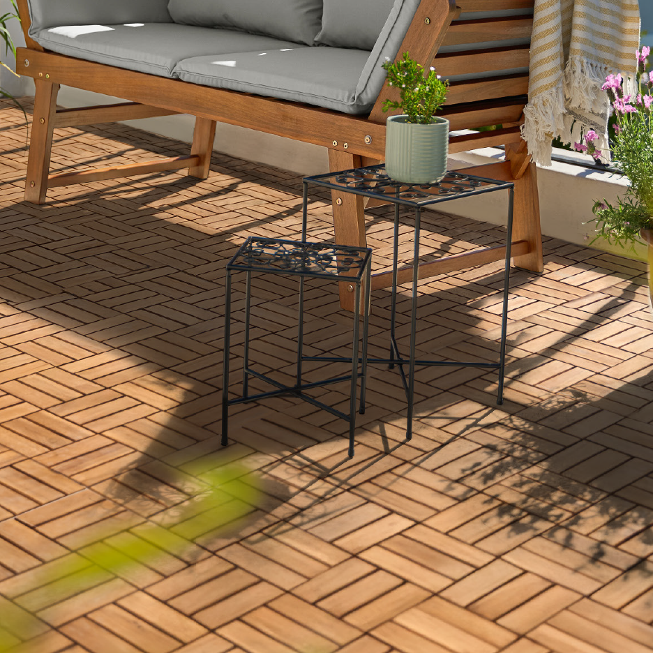 Trægulv på en balkon med en træbænk med grå hynder og to sorte metalborde med planter.