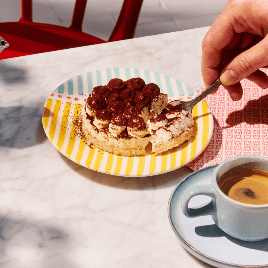 En hånd tager en skefuld tiramisu fra en farverig tallerken ved siden af en kop kaffe.