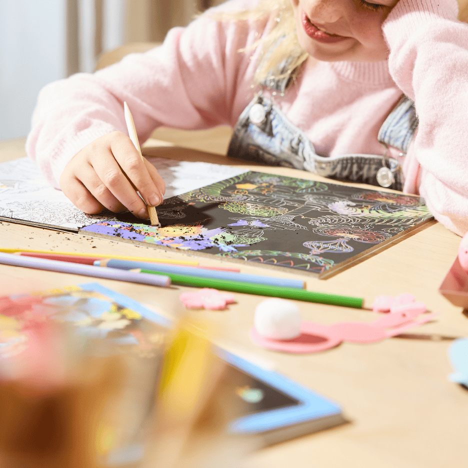 Et barn skraber et farverigt skrabelod med en træpind, omgivet af farveblyanter.