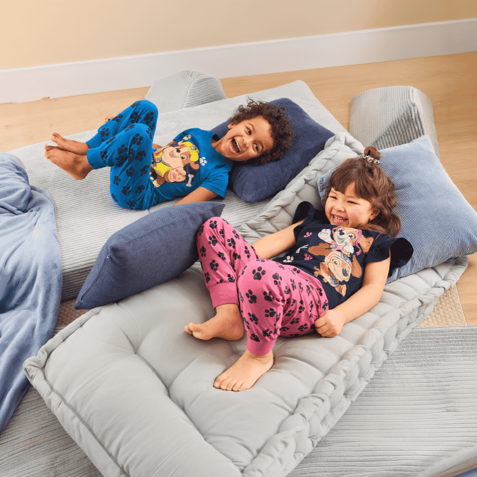 To glade børn i pyjamas ligger på grå gulvpuder og puder.