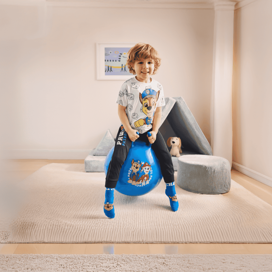 Glad dreng på en blå hoppebold med tegneseriefigurer, iført matchende t-shirt og sokker.