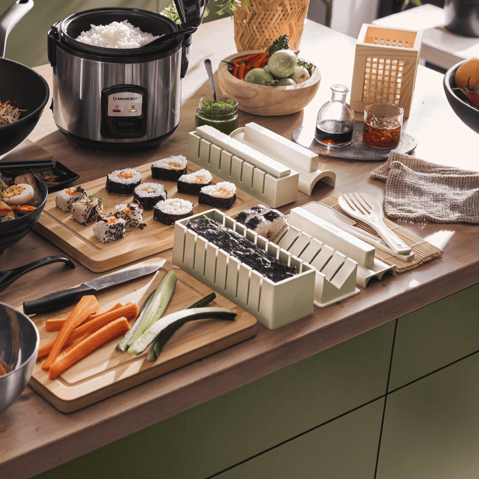 Rismaskine og sushisæt på et køkkenbord med frisklavet sushi og grøntsager.