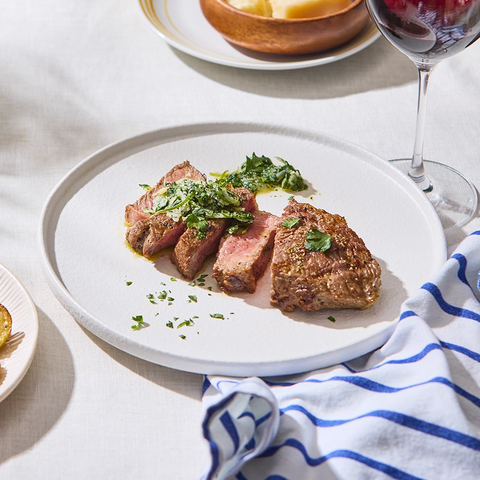 Skiver af grillet bøf med grøn sauce og persille på en hvid tallerken.
