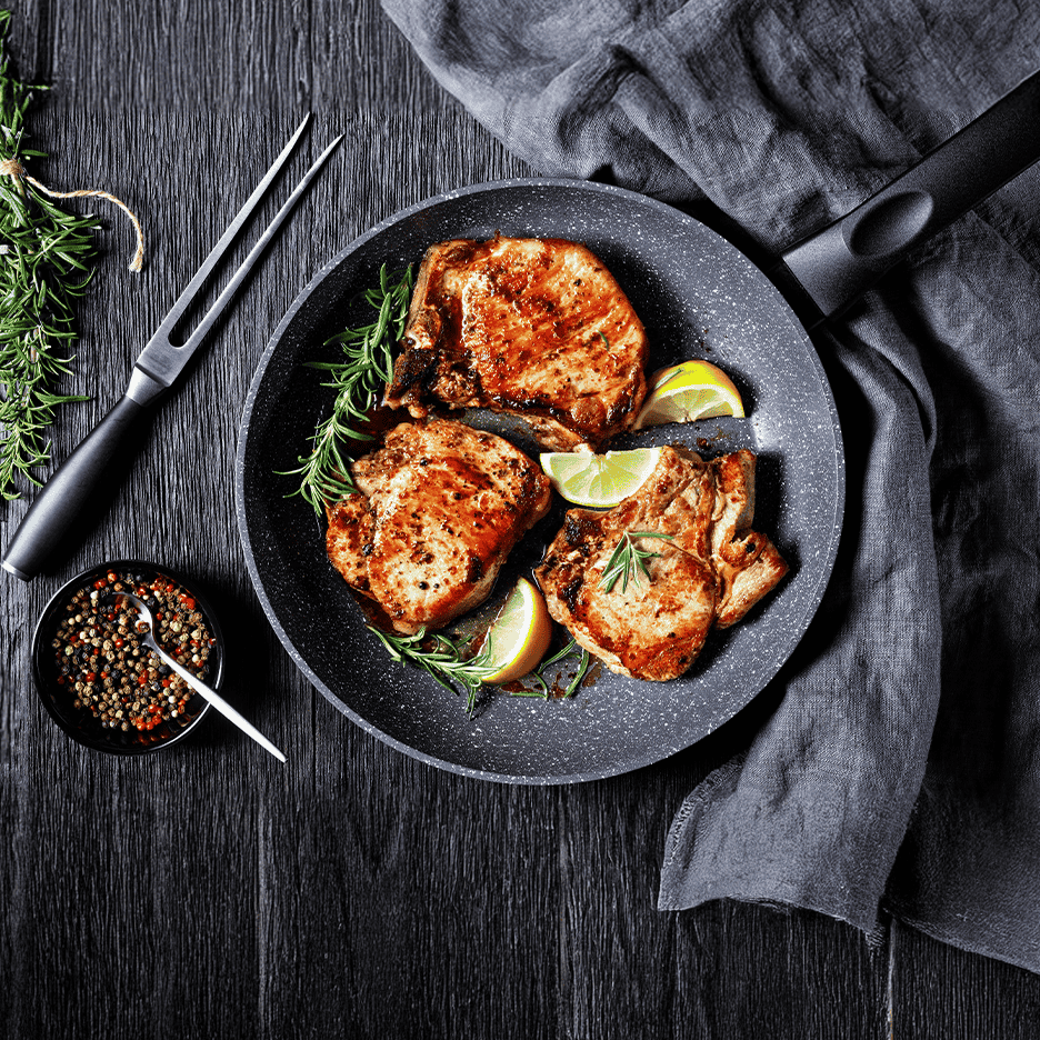 Stegte svinekoteletter med rosmarin og lime i en pande på et mørkt træbord.