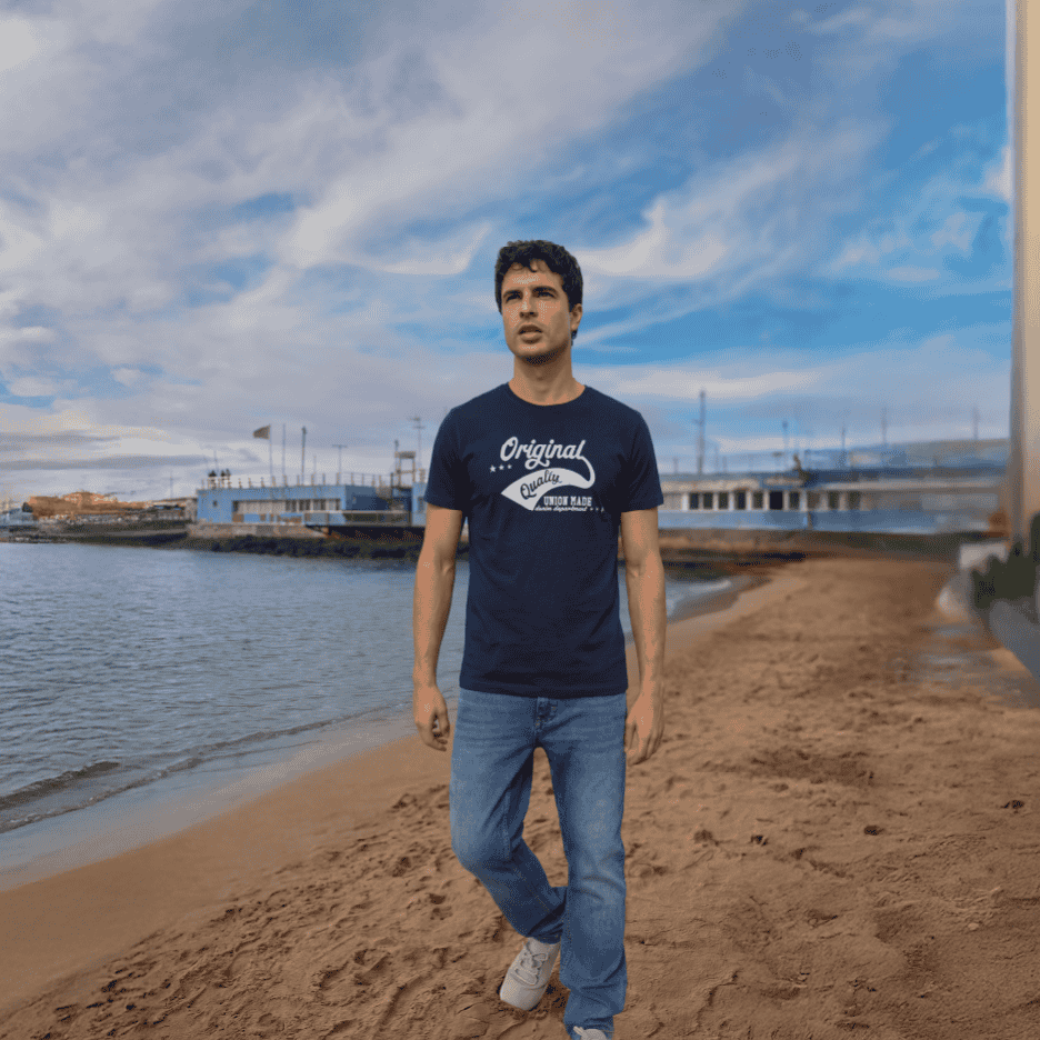 Mand i mørkeblå t-shirt med 'Original Quality' print og jeans går på stranden.