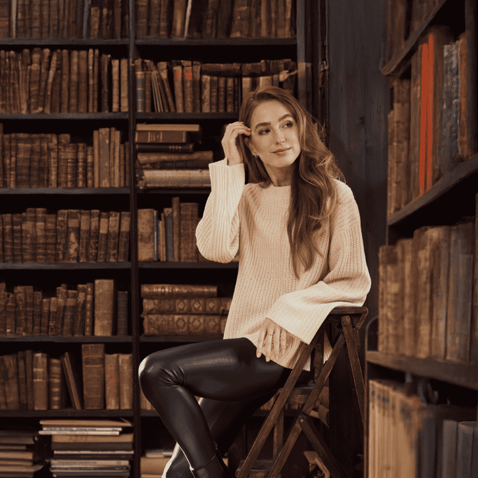 Kvinde i striktrøje og læderbukser sidder på en stige i et bibliotek.