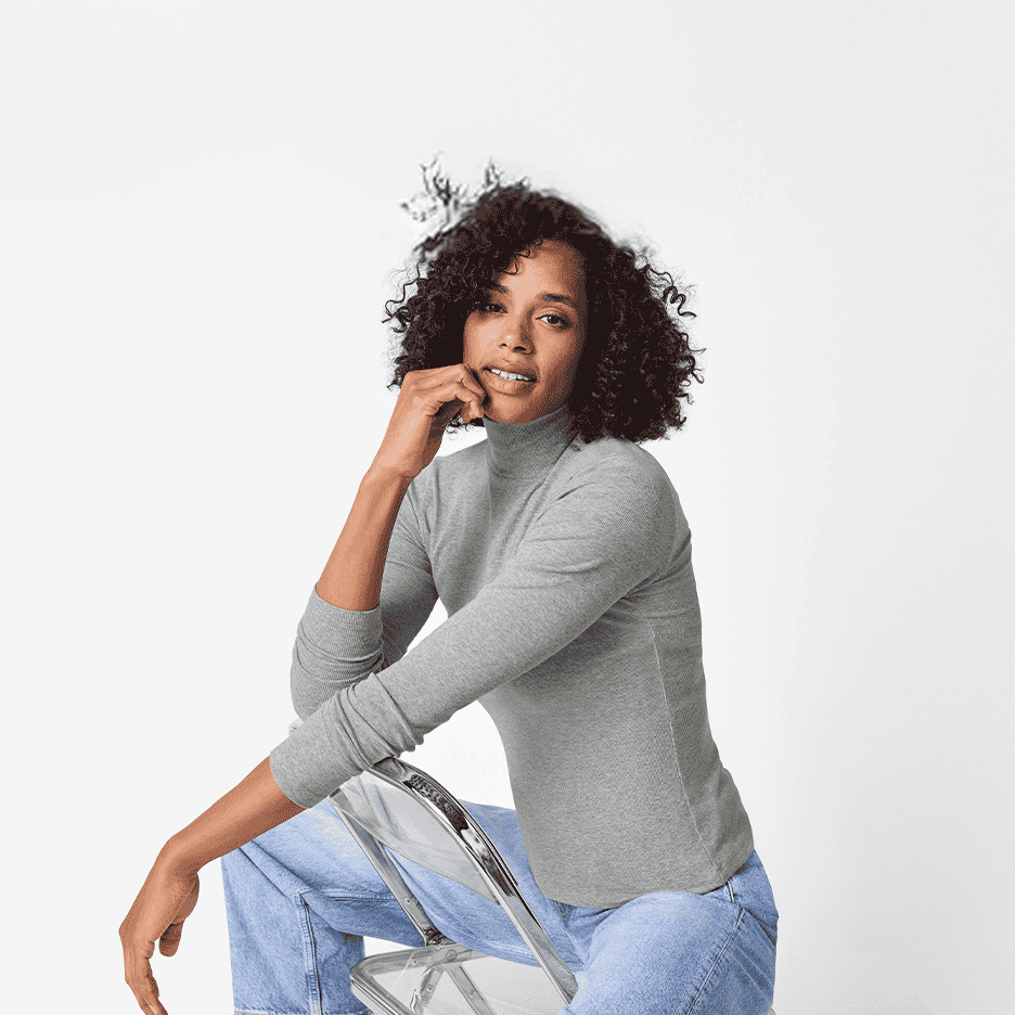 Kvinde i grå rullekrave og jeans sidder på en gennemsigtig stol.