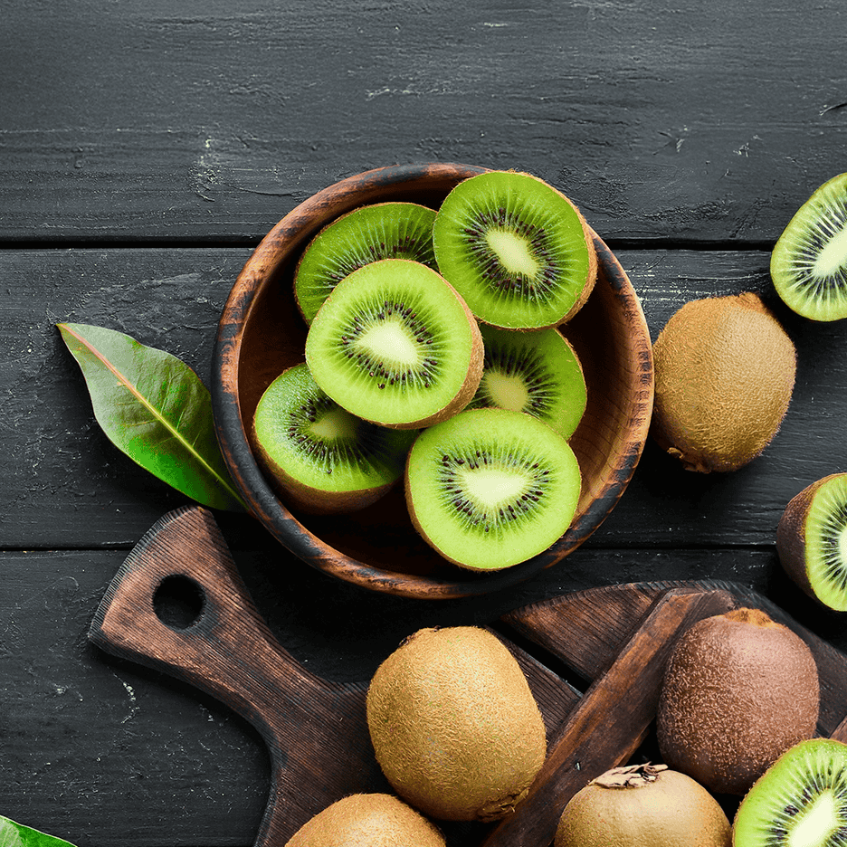 Skåret kiwi i en træskål og hele kiwifrugter på et mørkt træbord.