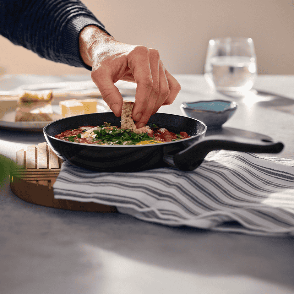 En person dypper brød i en pande med shakshuka, serveret på et bord med vandglas.