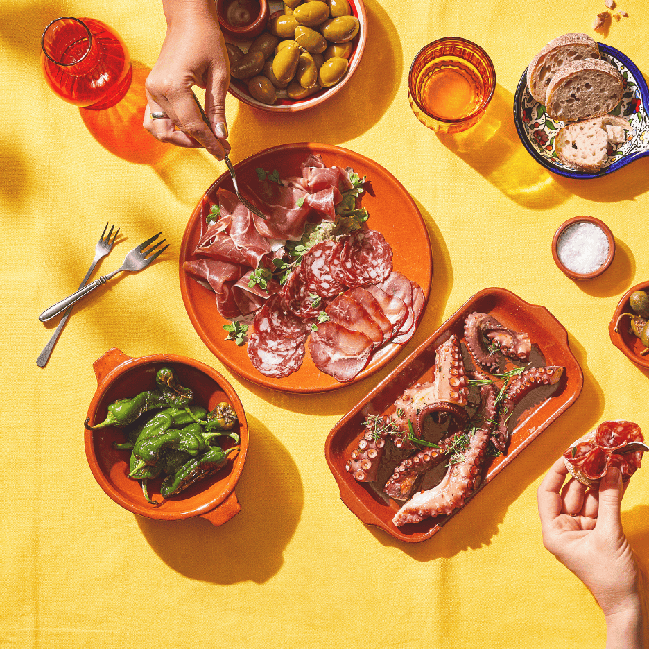 Tapasbord med charcuteri, blæksprutte, oliven, peberfrugter og brød på en gul dug.