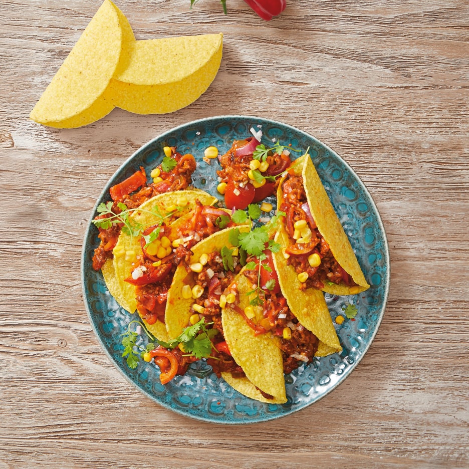 En tallerken med tacoskaller fyldt med hakket kød, majs, løg og peberfrugt, med ekstra tacoskaller ved siden af.