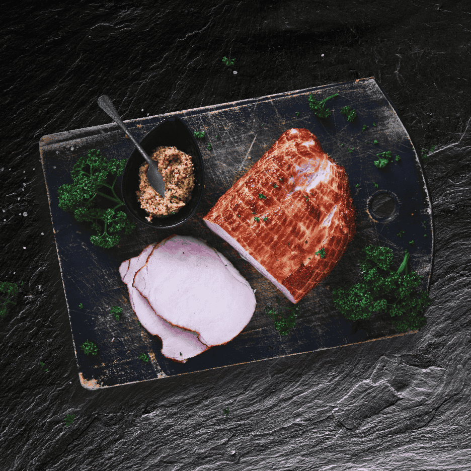Skåret svinekam med sennep og persille på et mørkt skærebræt.