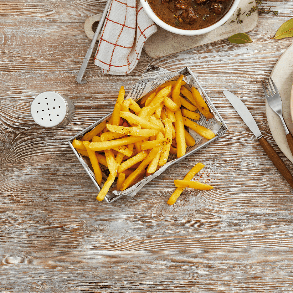 Pommes frites i en metalbakke med en skål gryderet og bestik på et træbord.
