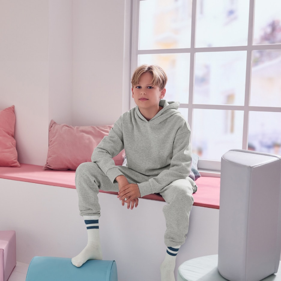 Dreng i grå hættetrøje og joggingbukser sidder i vindueskarm med lyserøde puder.
