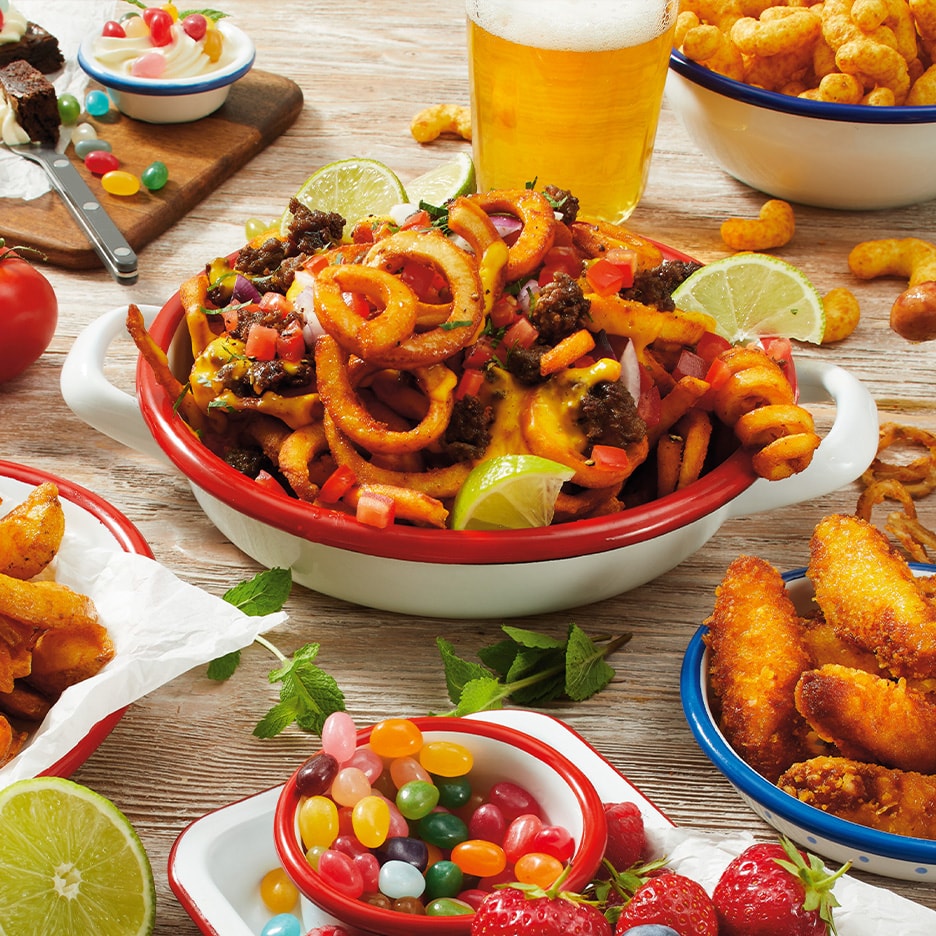 En skål med krøllede fritter toppet med hakket kød, ost og grøntsager, omgivet af snacks og slik.