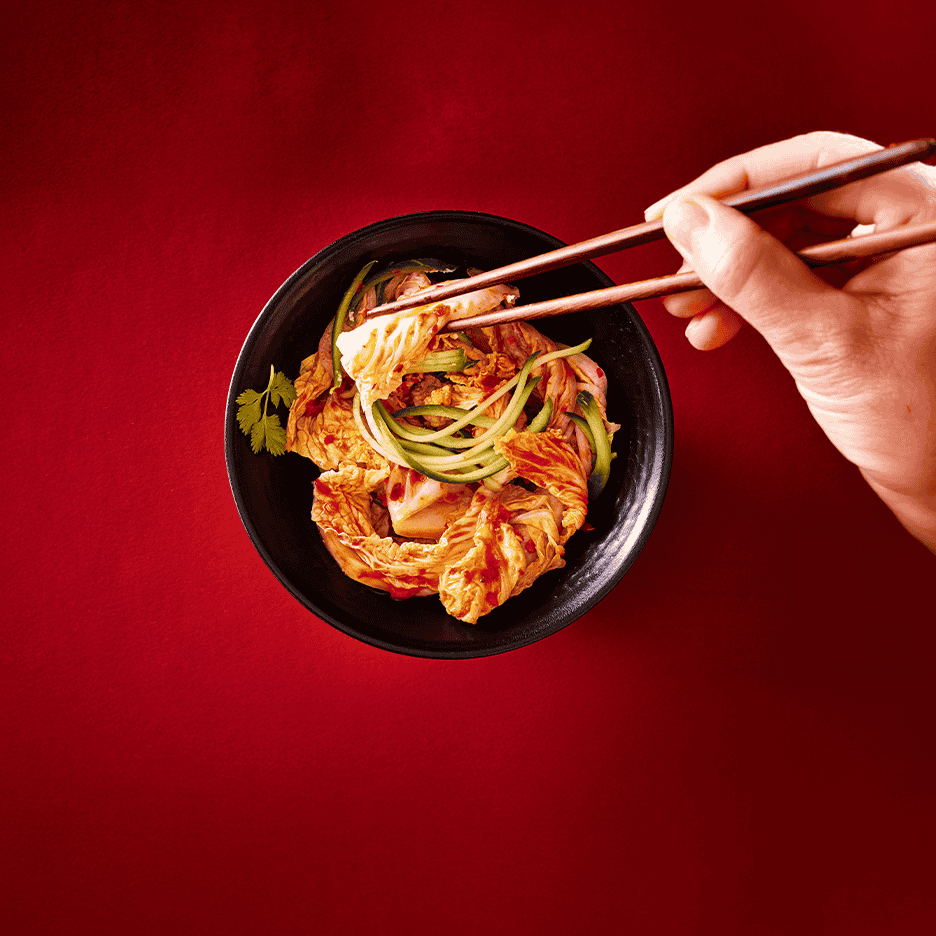 En skål med kimchi og agurkestrimler, der løftes med spisepinde på en rød baggrund.