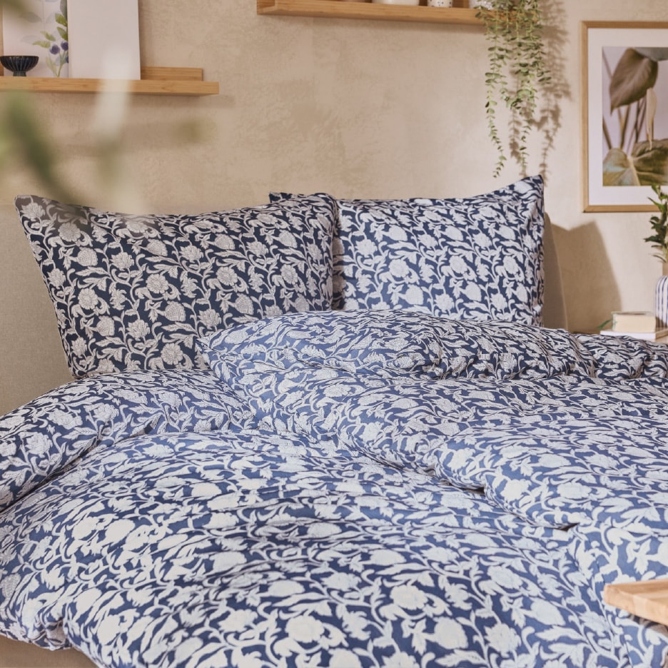 Sengesæt med blå og hvidt blomstermønster på en seng med træhylder og planter i baggrunden.