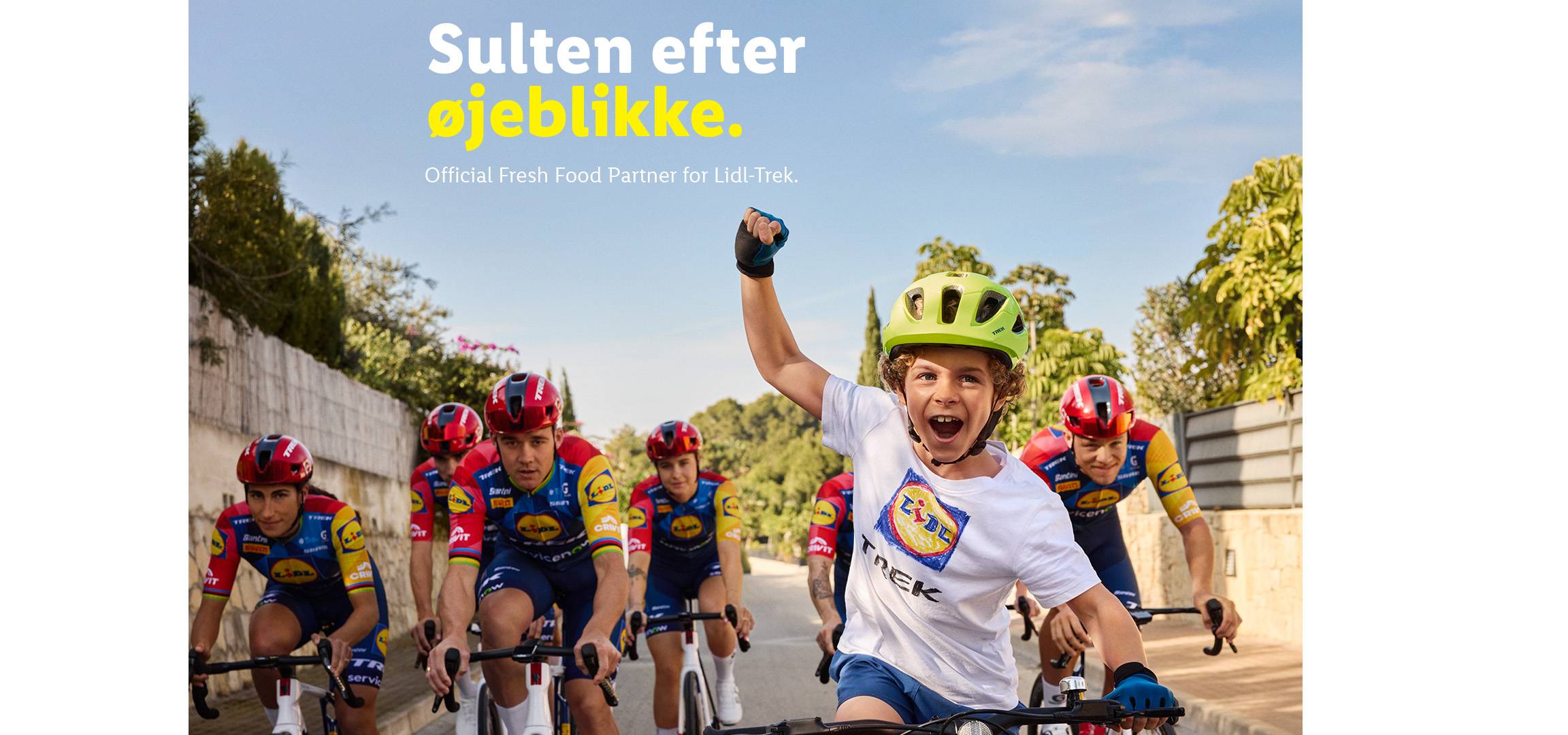 En dreng i en grøn cykelhjelm jubler, mens cykelryttere i farverige trøjer cykler bag ham.